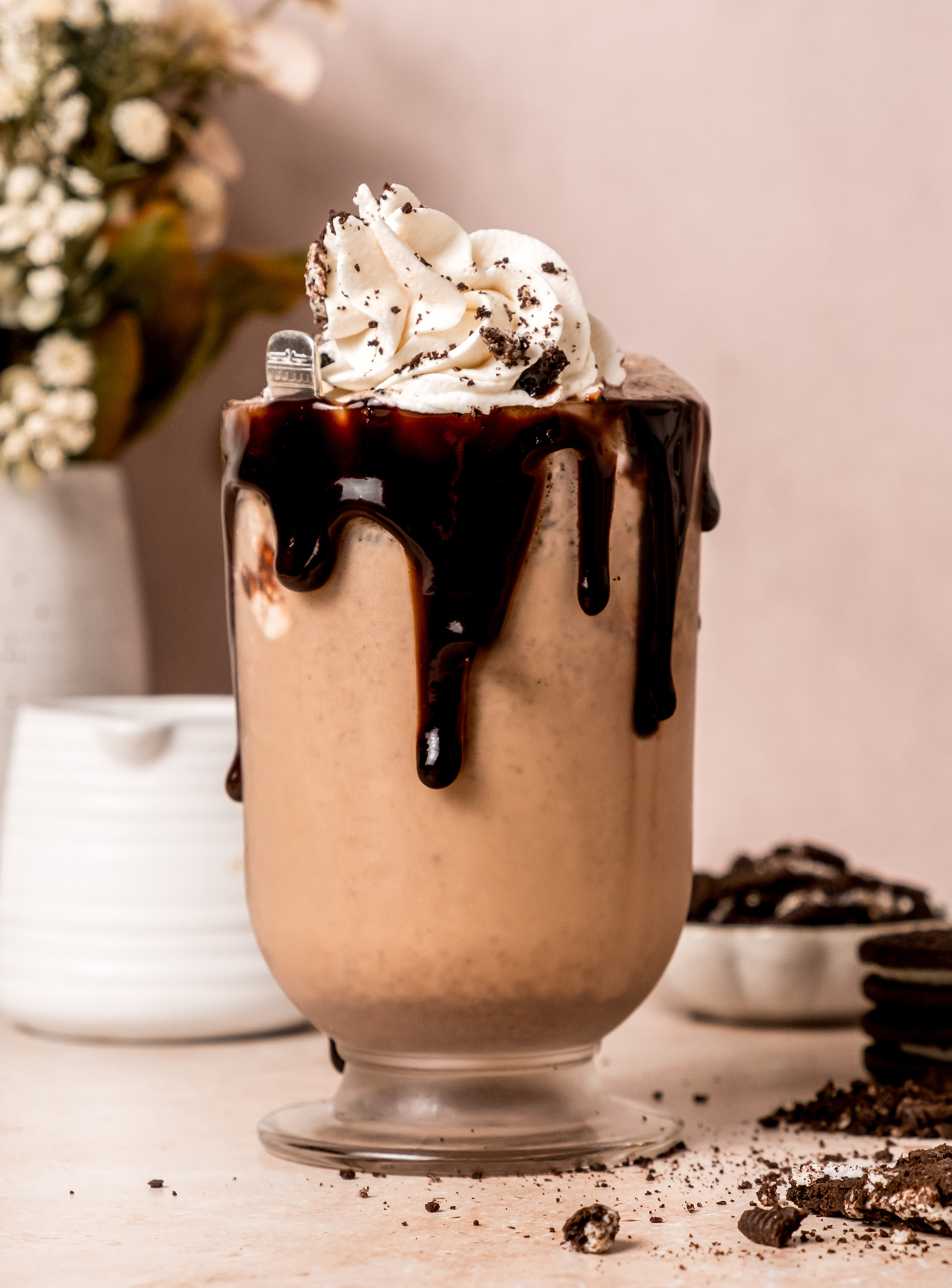 A tall milkshake glass filled with blended cookies and cream milkshake, topped with whipped cream and cookie bits, with a flower vase and more cookies in the background.