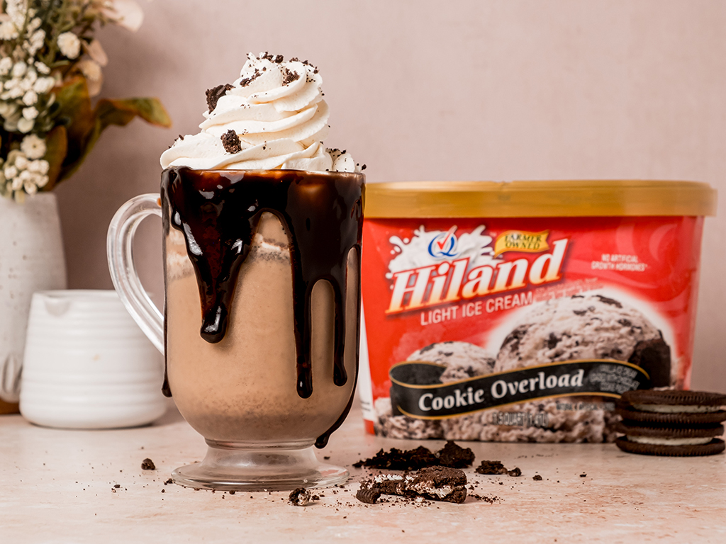 A cookies and cream milkshake served in a clear glass mug with chocolate sauce dripping over the sides, topped with whipped cream and cookie pieces. A container of Hiland Cookie Overload Ice Cream is behind the milkshake alongside crumbled cookies.