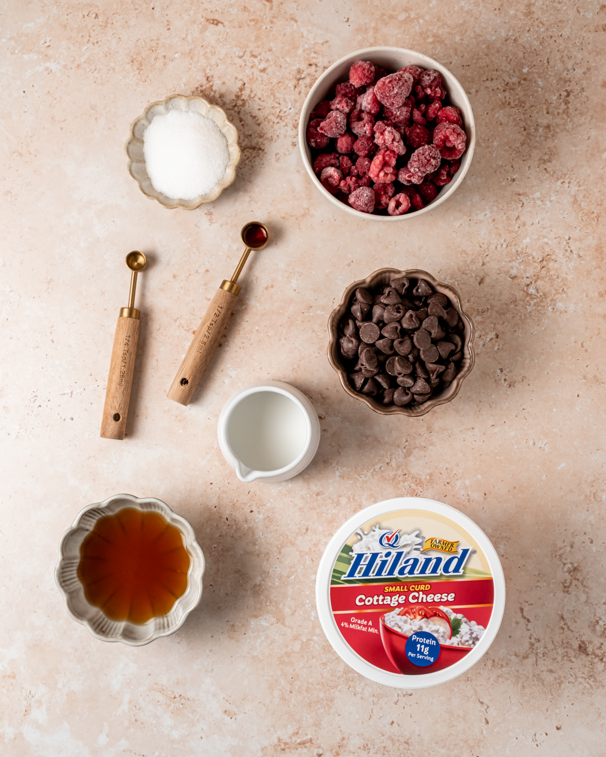 A flat-lay of ingredients for cottage cheese ice cream, including frozen raspberries, chocolate chips, a container of Hiland cottage cheese, sugar, vanilla extract, maple syrup, and a small bowl of milk.