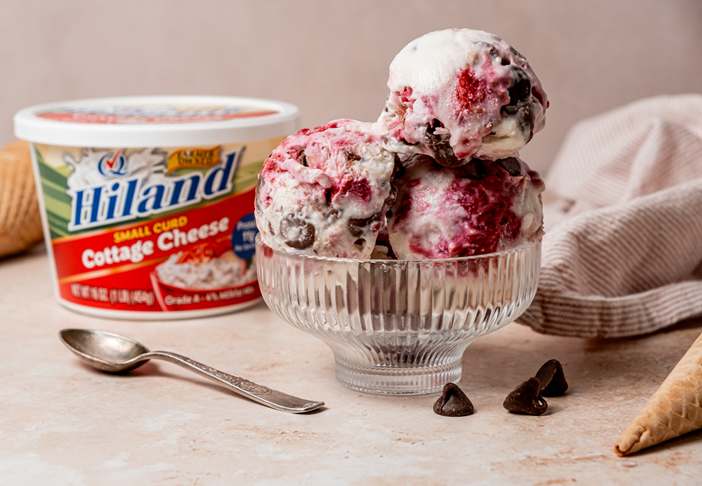 A close-up of a clear glass dessert bowl filled with three scoops of cottage cheese ice cream swirled with bright raspberry and studded with dark chocolate chips, placed in front of a tub of Hiland Small Curd Cottage Cheese.