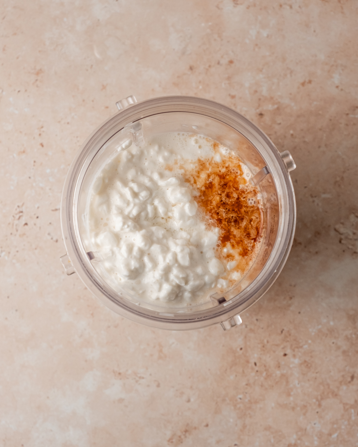 A blender cup filled with cottage cheese, vanilla, and maple syrup before blending, set against a light tan surface.