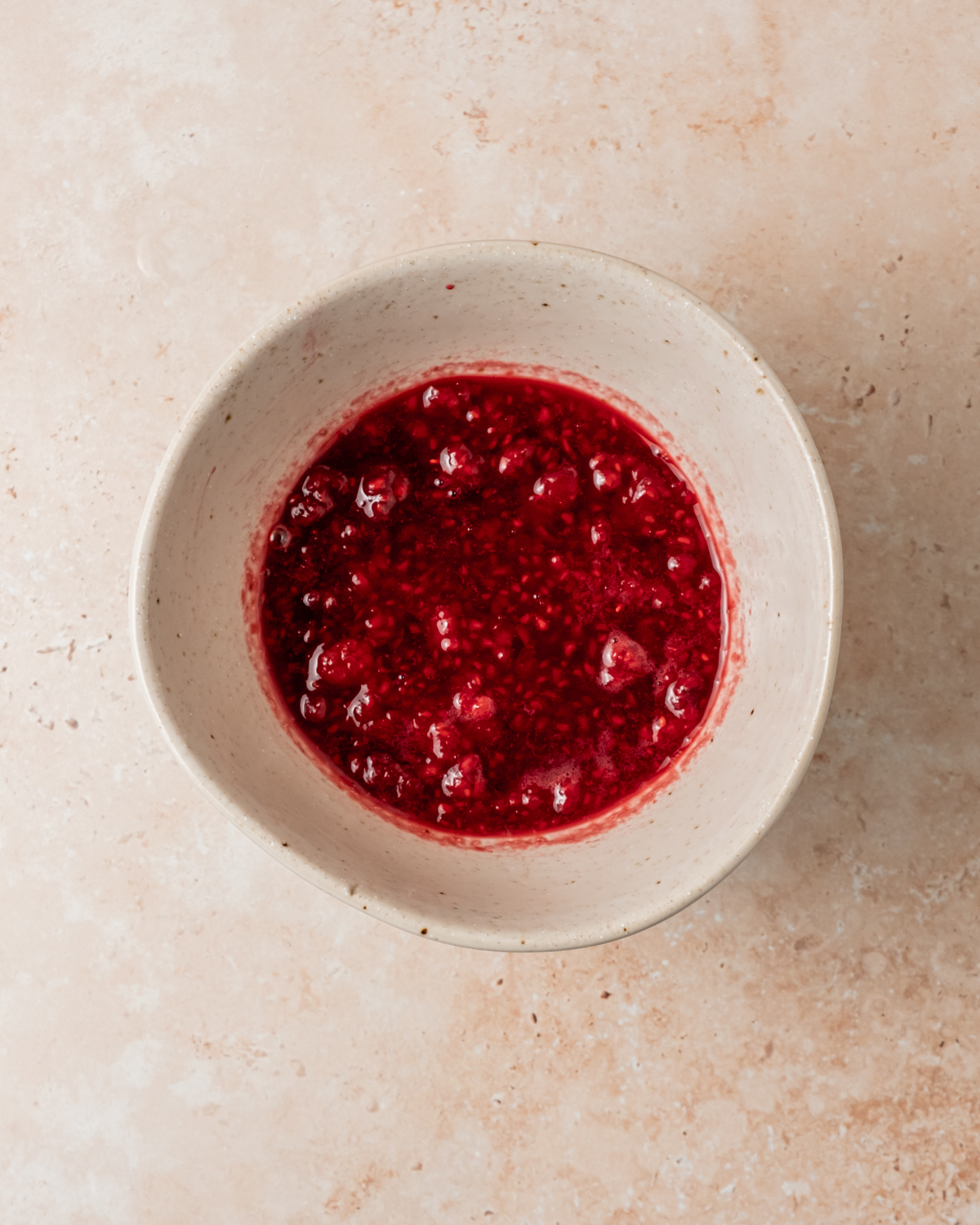 A bowl filled with a mashed raspberry mixture showing a bright red hue and juicy texture on a beige countertop.