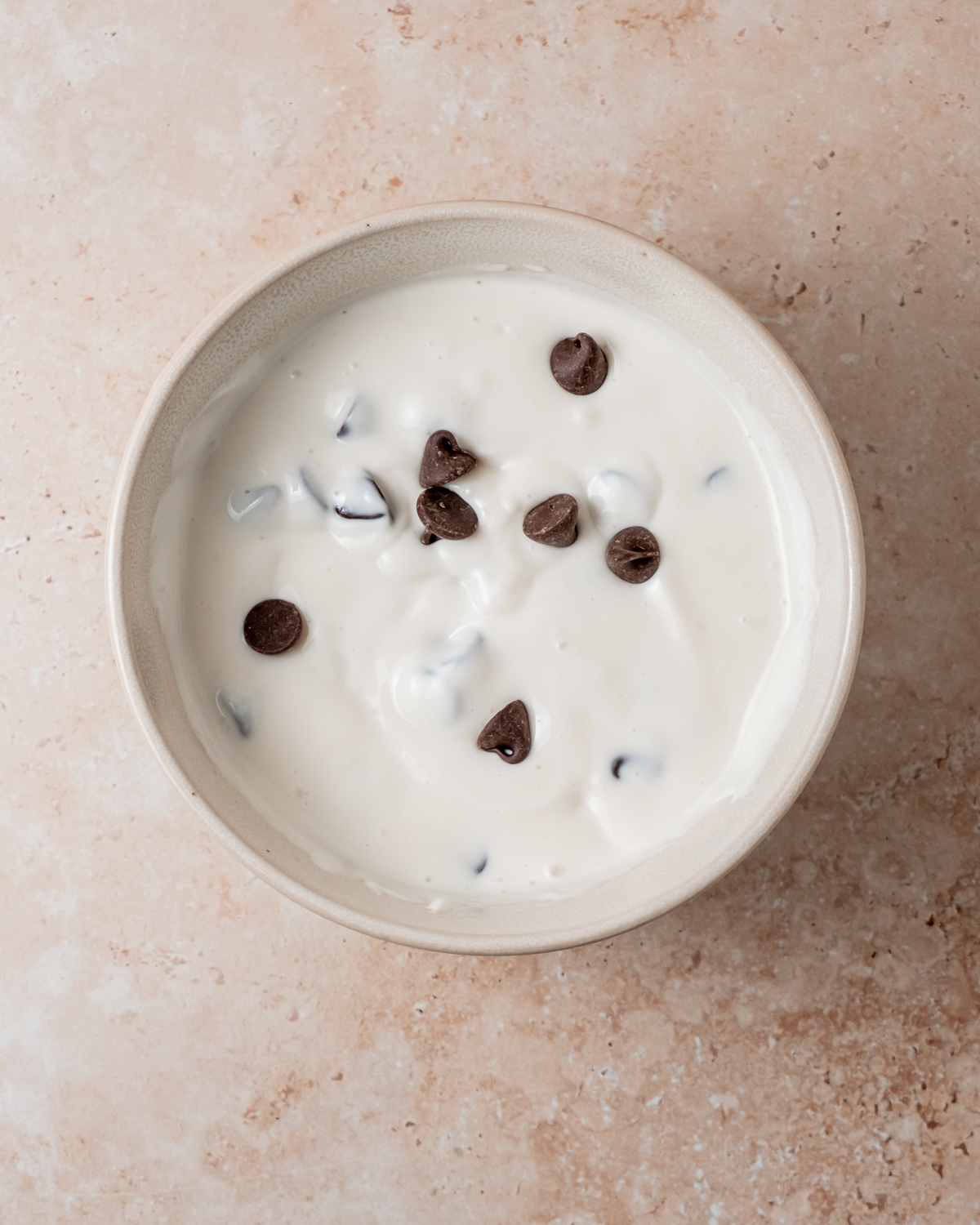 A beige ceramic bowl containing a creamy cottage cheese mixture blended with chocolate chips, sitting on a light tan countertop.