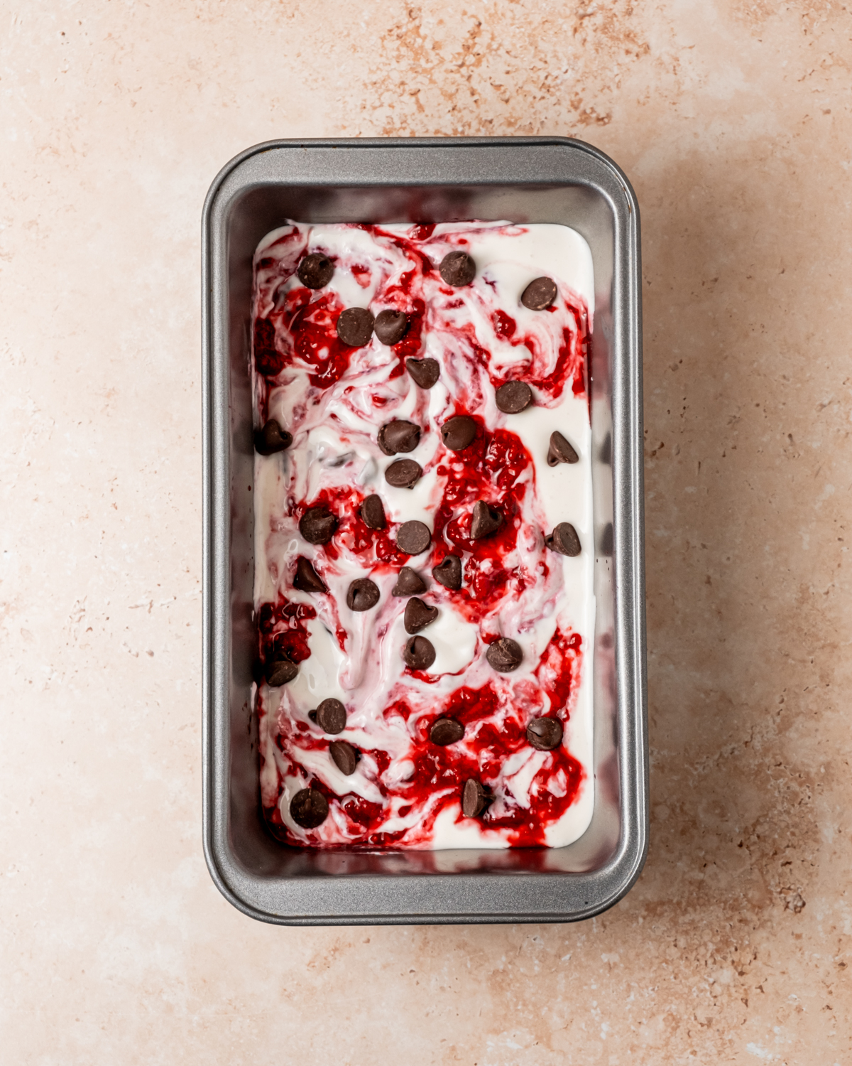 A rectangular metal pan filled with a creamy mixture swirled with bright red raspberry sauce and sprinkled with chocolate chips, ready to be frozen into cottage cheese ice cream.