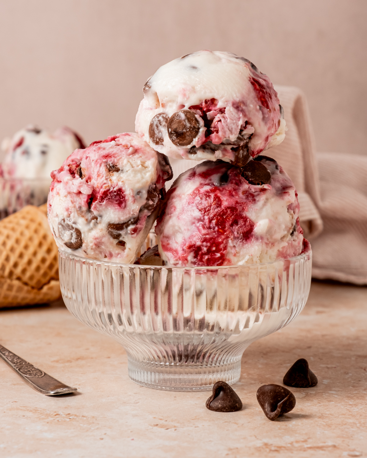 A slightly angled view of three scoops of raspberry chocolate chip cottage cheese ice cream in a ribbed glass bowl, with chocolate chips scattered nearby and waffle cones and a striped napkin in the background.