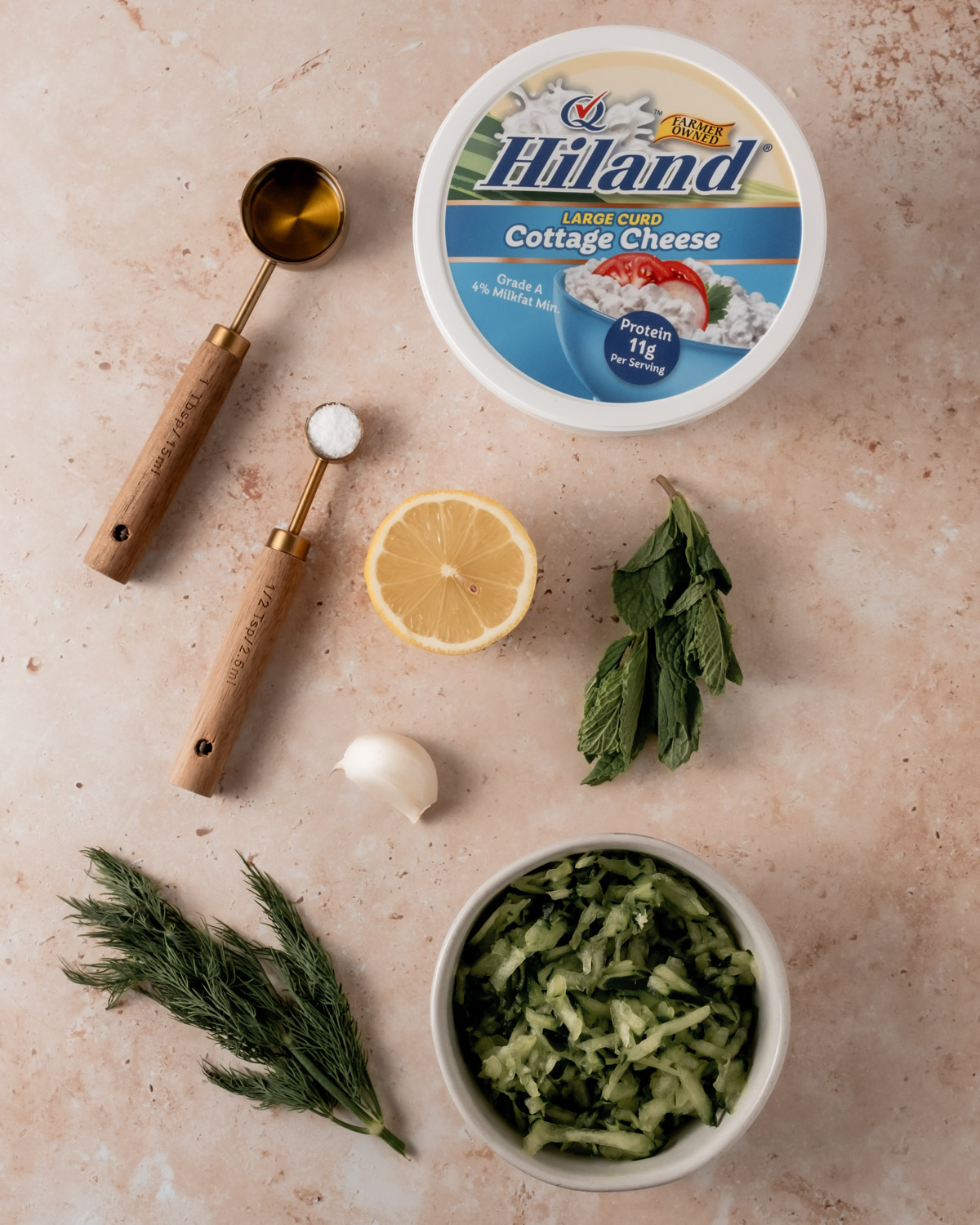 A close-up of tzatziki dip ingredients arranged on a beige background, including a tub of Hiland Large Curd Cottage Cheese, fresh dill, mint, grated cucumber, lemon, garlic, and measuring spoons with salt and oil.