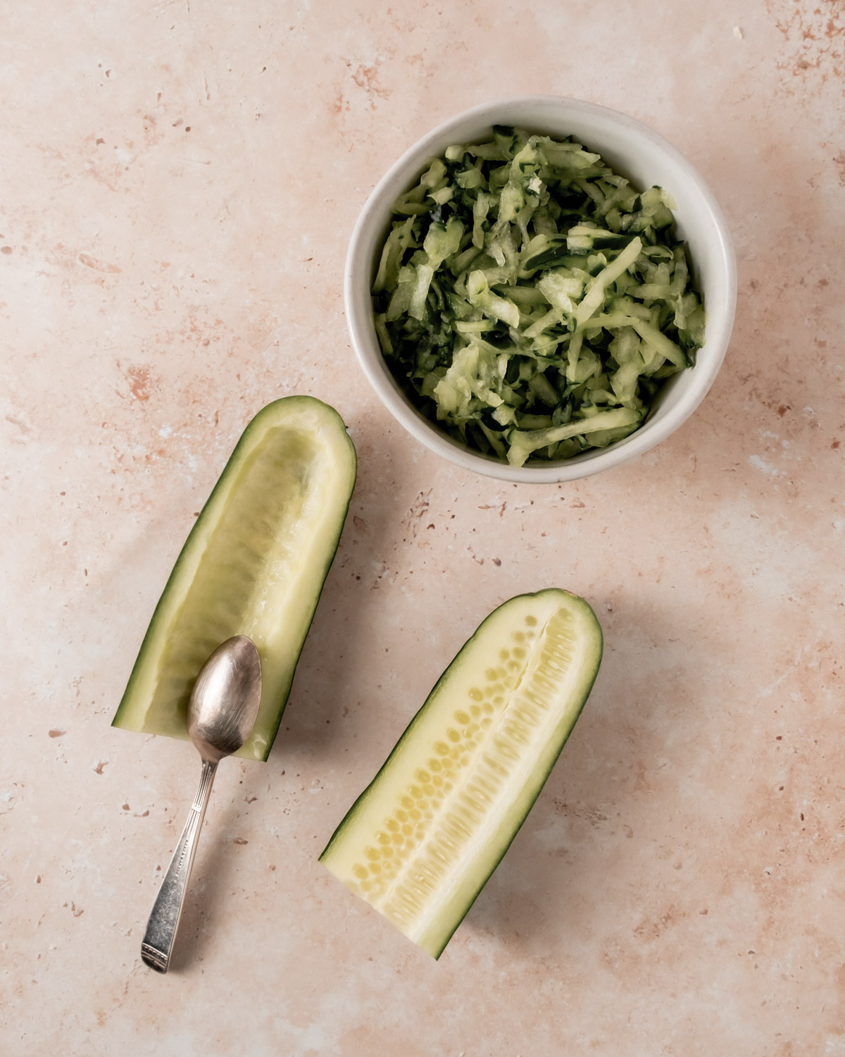 A bowl of grated cucumber with a halved cucumber beside it, showing the scooped-out seeds. A small silver spoon rests on the vegetable.