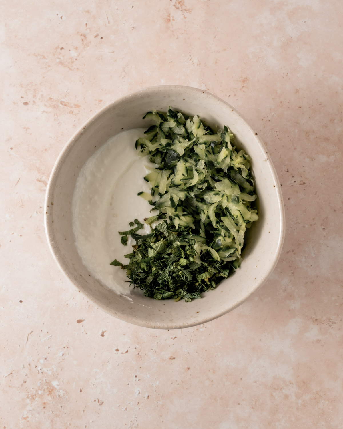 A mixing bowl with cottage cheese and finely chopped herbs, including dill and mint, partially mixed into the base of the tzatziki dip.