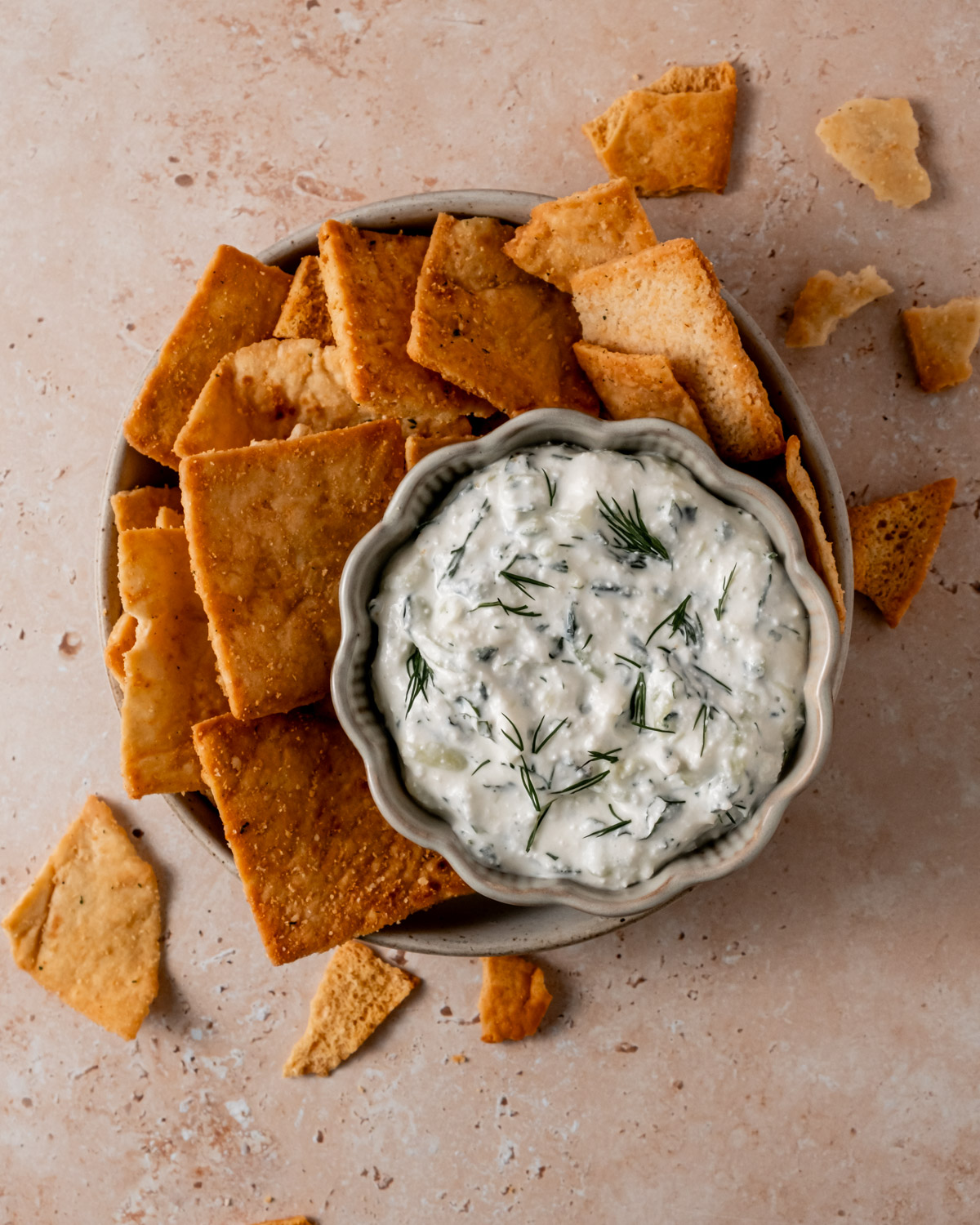 A bowl of creamy cottage cheese tzatziki dip garnished with fresh dill, surrounded by crispy pita chips on a neutral-toned surface. The dip has a thick texture with visible herbs and cucumber pieces.