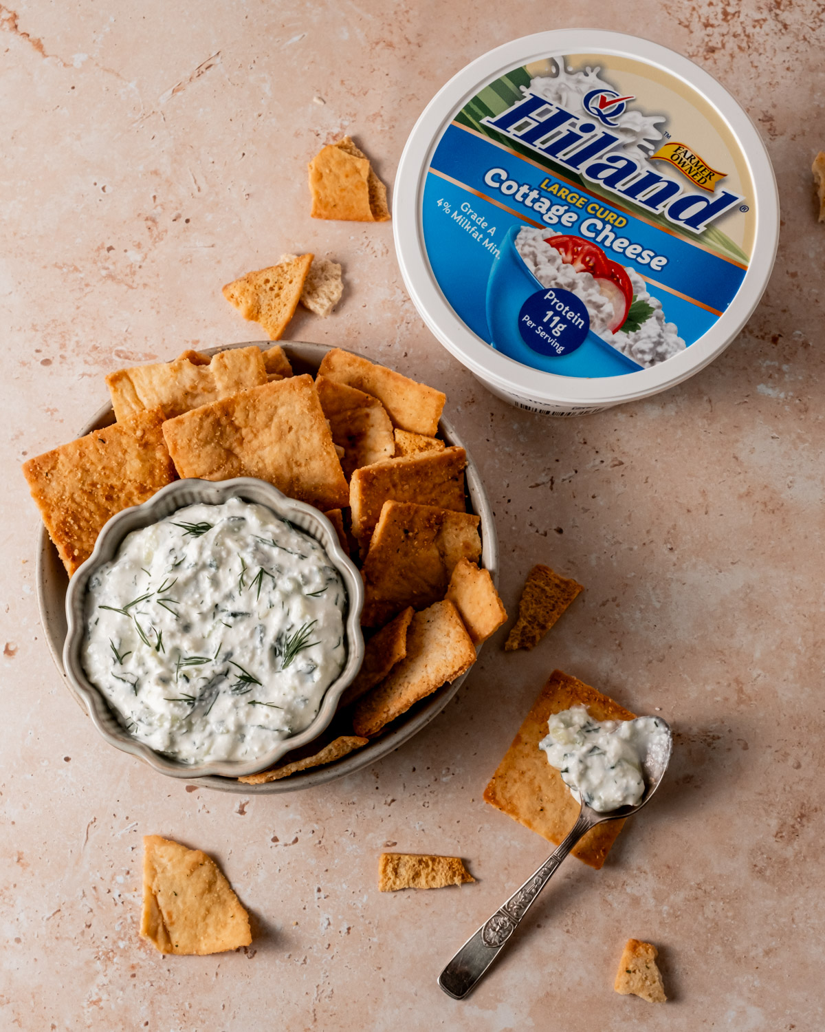 A bowl of creamy cottage cheese tzatziki dip garnished with fresh dill, surrounded by crispy pita chips on a textured beige surface. A container of Hiland Large Curd Cottage Cheese sits beside the bowl, emphasizing the key ingredient.