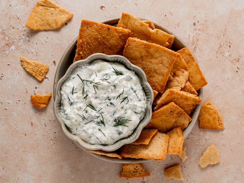 A close-up of a small bowl filled with creamy tzatziki dip made with cottage cheese, garnished with fresh dill, and surrounded by golden pita chips. Broken chip pieces are scattered around for a rustic touch.