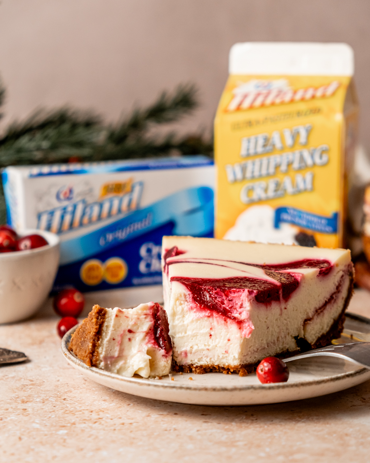 A slice of cranberry white chocolate cheesecake on a small plate, with a swirl of red cranberry sauce over creamy filling and a graham cracker crust, surrounded by fresh cranberries. Behind, a Hiland Cream Cheese package and Hiland Heavy Whipping Cream carton are visible.