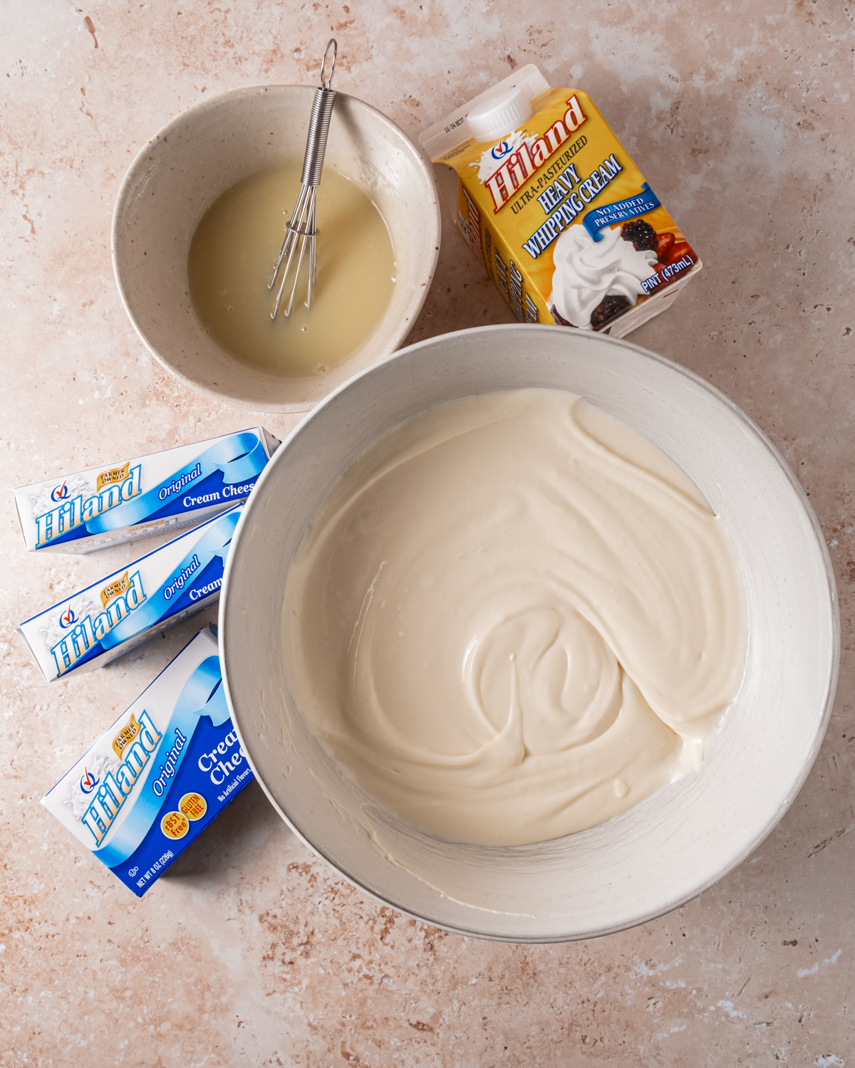 Ingredients for cranberry white chocolate cheesecake displayed on a countertop. Ingredients include Hiland cream cheese, heavy whipping cream, white chocolate, eggs, sugar, unsalted butter, and other baking essentials.