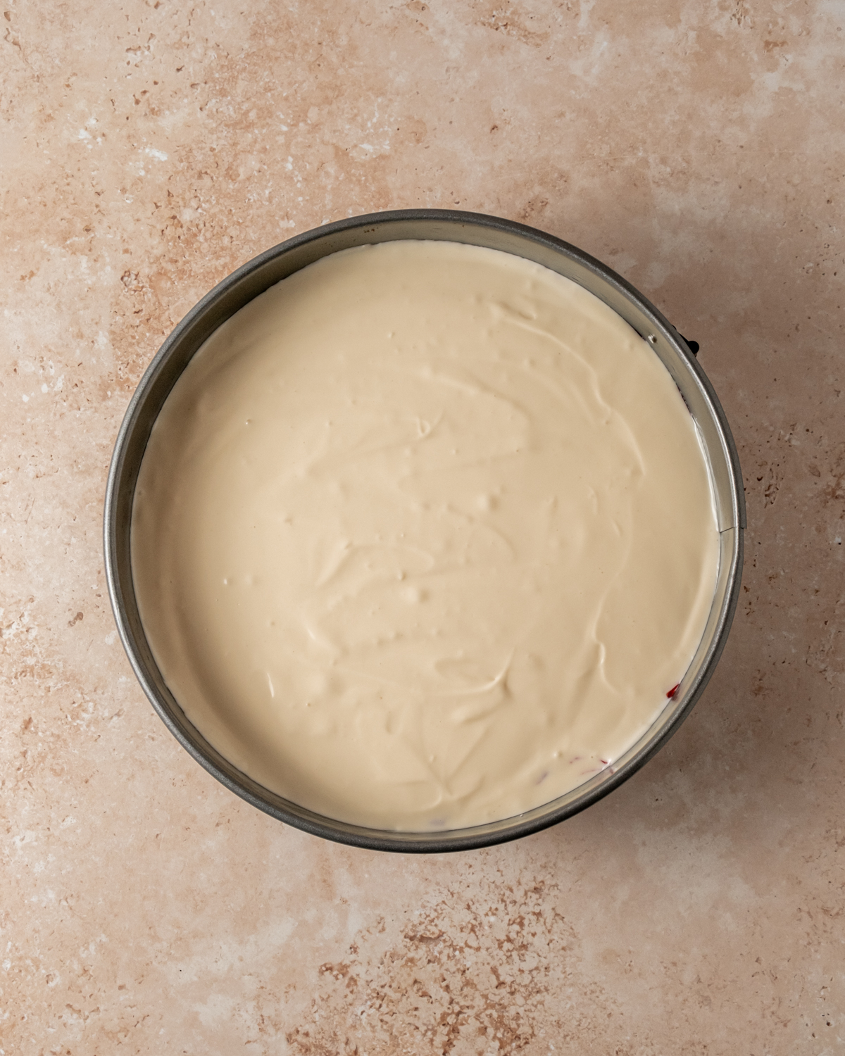 A smooth layer of creamy cheesecake filling poured into a springform pan over the prepared crust, ready for baking. The filling has an even, glossy texture.