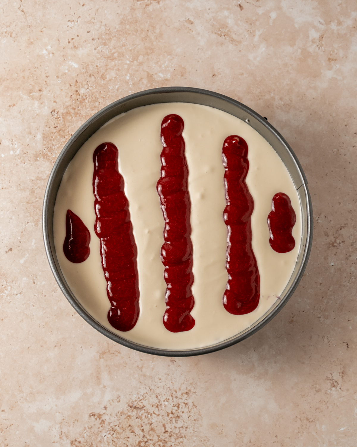 A springform pan filled with creamy cheesecake batter topped with five vertical stripes of vibrant cranberry sauce, ready to be swirled for a marbled effect. The deep red contrast against the pale cheesecake filling adds a festive look.