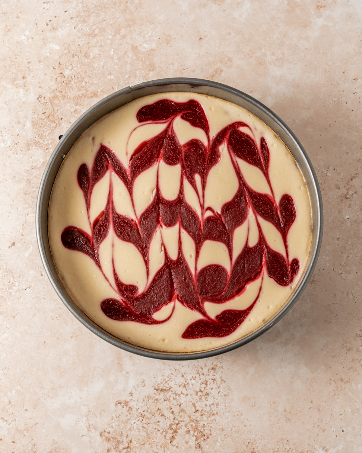 A close-up of a cheesecake in a springform pan with a baked cranberry swirl pattern. The cranberry design remains vibrant and intact, now set into the cheesecake’s surface for a visually striking presentation against the creamy, pale background.