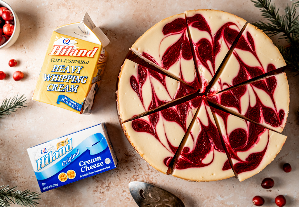 Top-down view of a cranberry white chocolate cheesecake, sliced into portions with a red cranberry swirl pattern over the surface. Hiland Heavy Whipping Cream and Hiland Cream Cheese packaging are positioned nearby, accompanied by scattered cranberries and greenery.