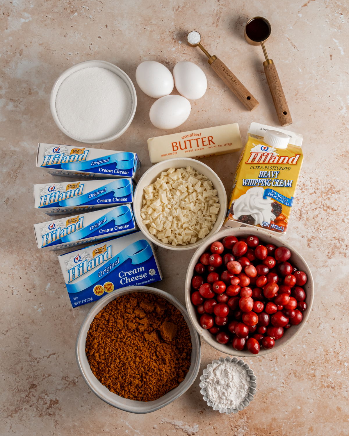 An assortment of cheesecake ingredients arranged on a light marble surface, including Hiland cream cheese blocks, heavy whipping cream, butter, cranberries, sugar, eggs, and other necessary ingredients for a cranberry cheesecake.