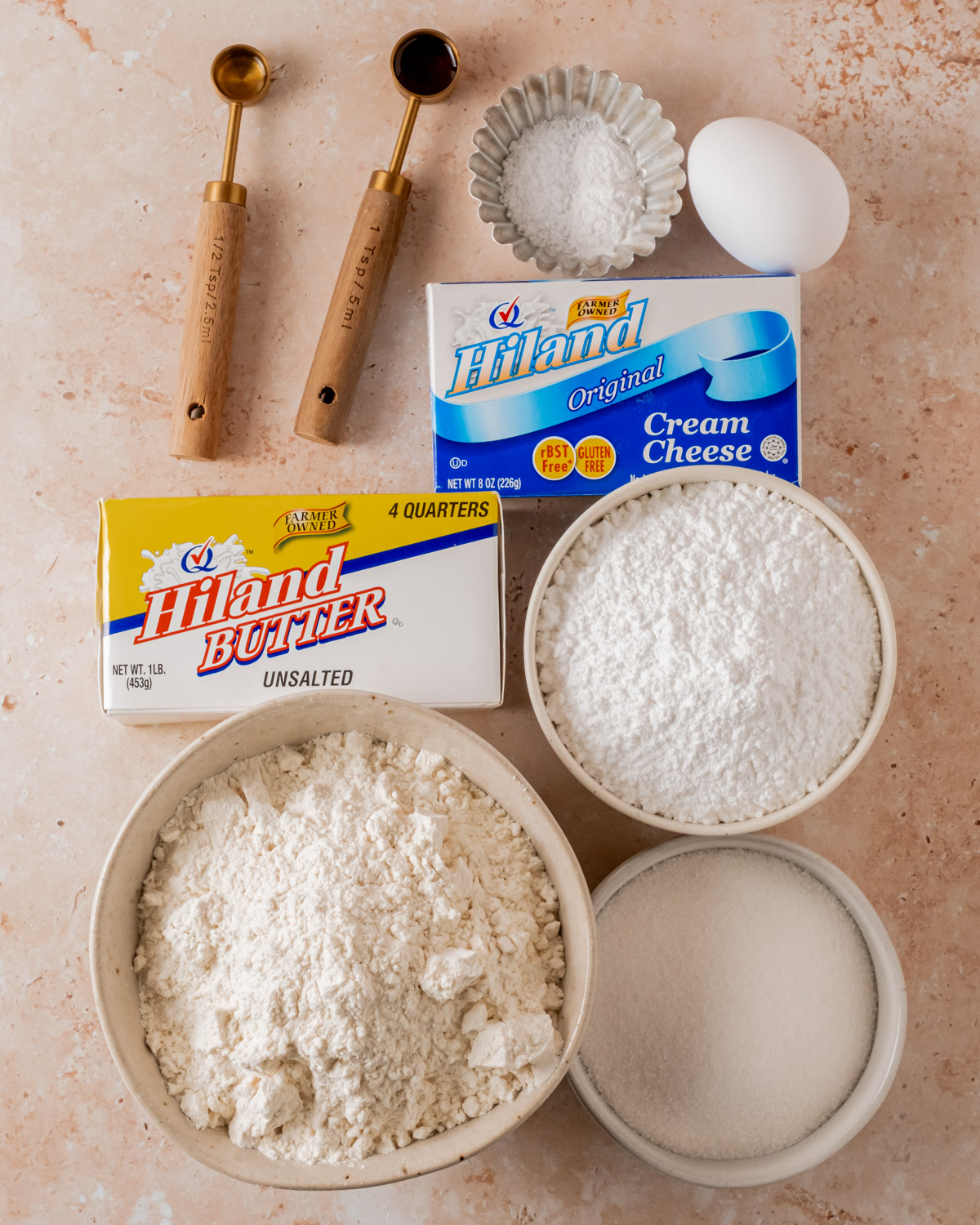 Ingredients for cream cheese cut-out cookies, including flour, sugar, butter, cream cheese, vanilla extract, and an egg, are arranged on a beige surface with wooden measuring spoons.