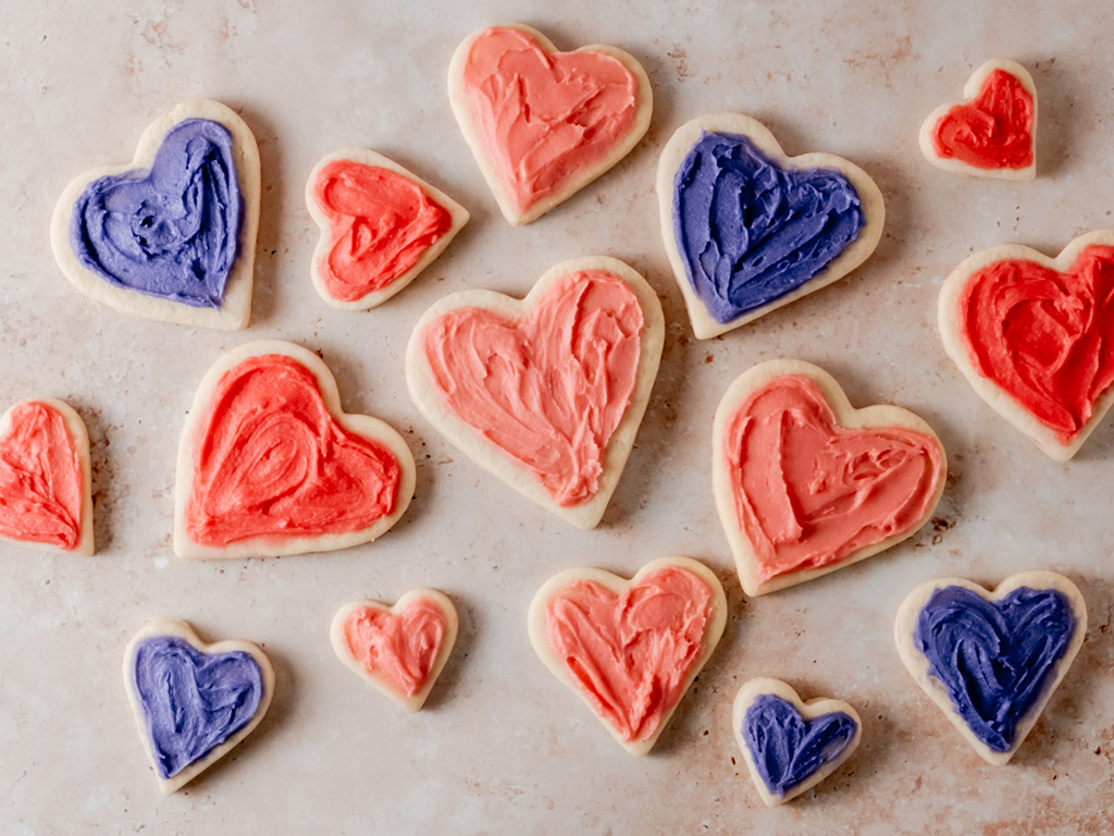 Heart-shaped sugar cookies in various sizes with pink and purple frosting, placed on a textured surface.