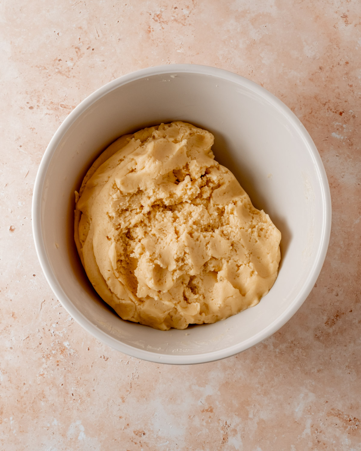 A bowl of soft, golden cookie dough sits in a white mixing bowl on a textured beige surface.