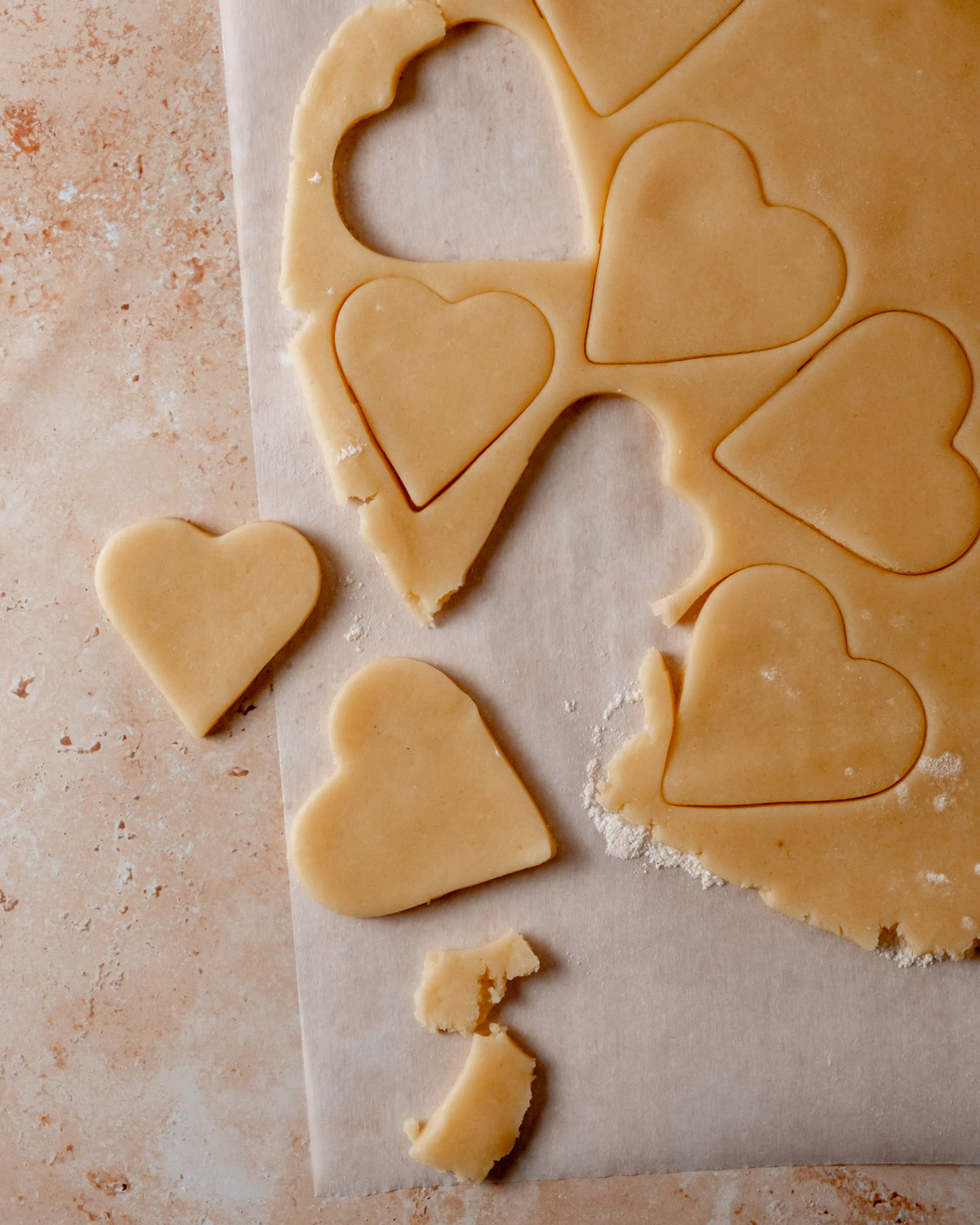 Heart-shaped cookie dough pieces are freshly cut from a rolled-out sheet, with some hearts still attached to the dough.