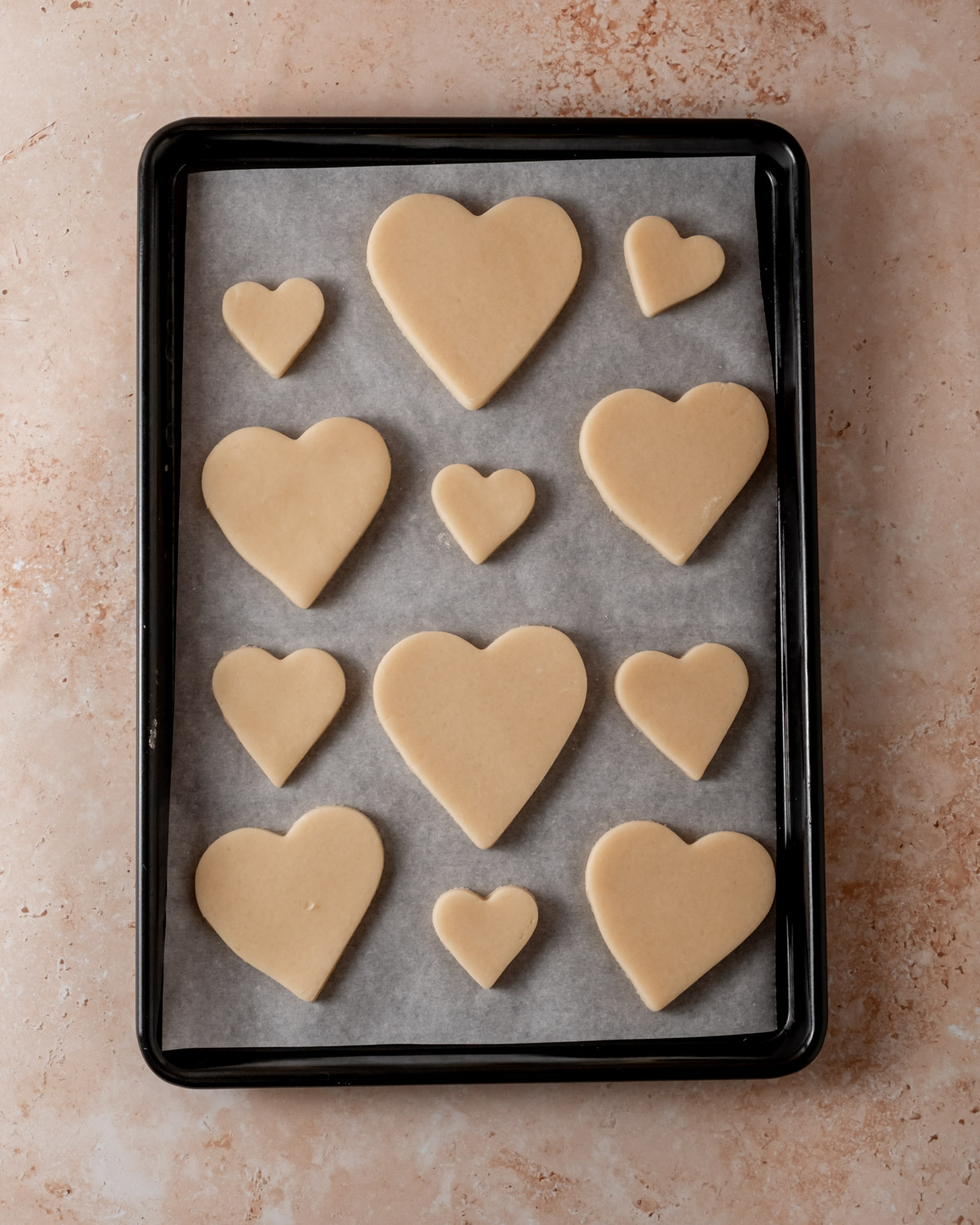 A black baking tray holds heart-shaped cookie dough cutouts arranged on parchment paper before baking.