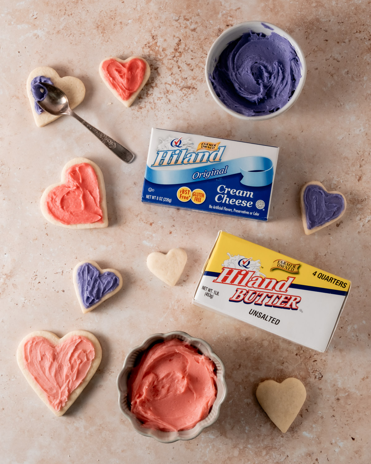 Heart-shaped sugar cookies are decorated with red and purple frosting, with a spoon resting in a bowl of frosting alongside butter and cream cheese packaging.