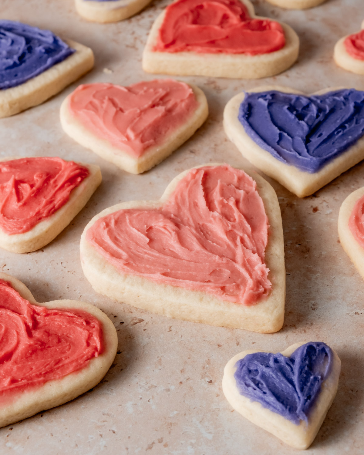 A close-up of heart-shaped sugar cookies with pink and purple frosting, arranged on a textured surface.