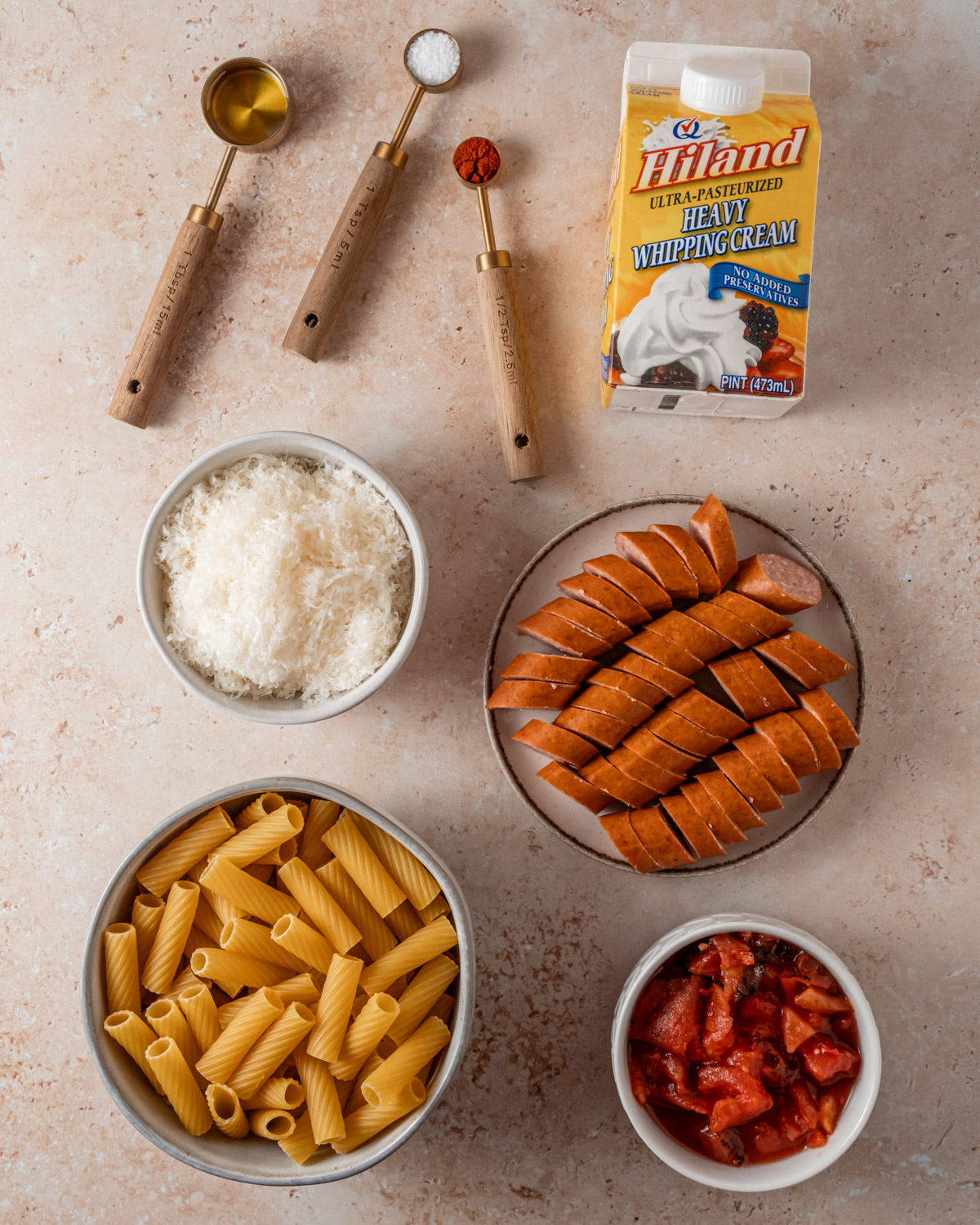 Ingredients for creamy sausage pasta are arranged on a surface, including rigatoni, grated Parmesan, sliced sausage, canned tomatoes, spices, and Hiland Heavy Whipping Cream.