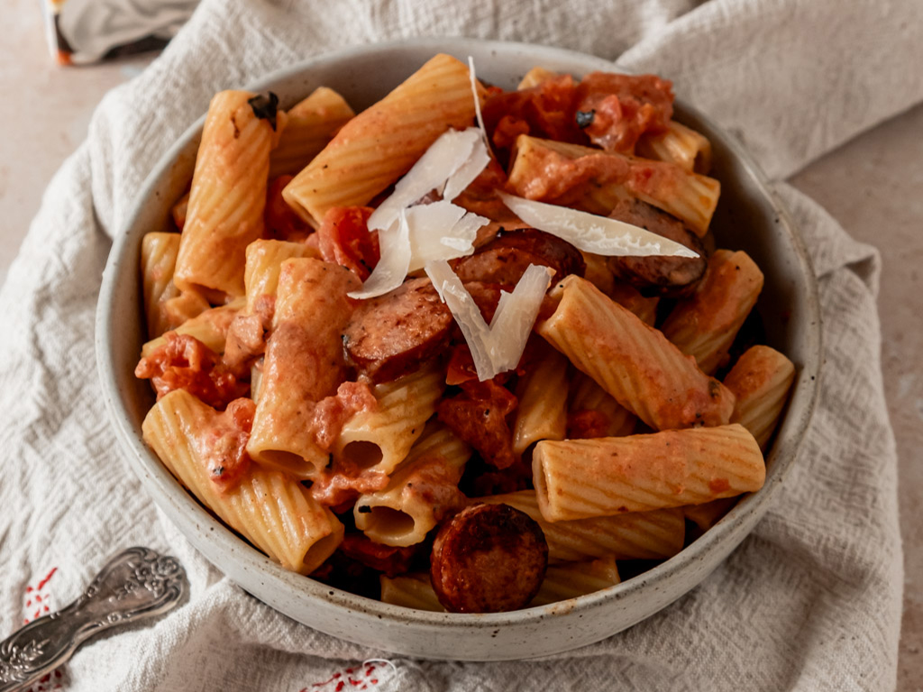 Close-up of creamy rigatoni pasta with sausage, tomato sauce, and Parmesan shavings in a rustic bowl.