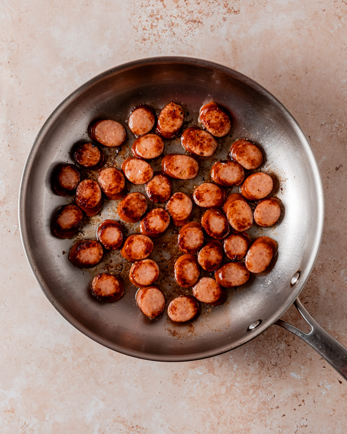 Slices of browned sausage are sautéed in a stainless steel skillet with caramelized edges.