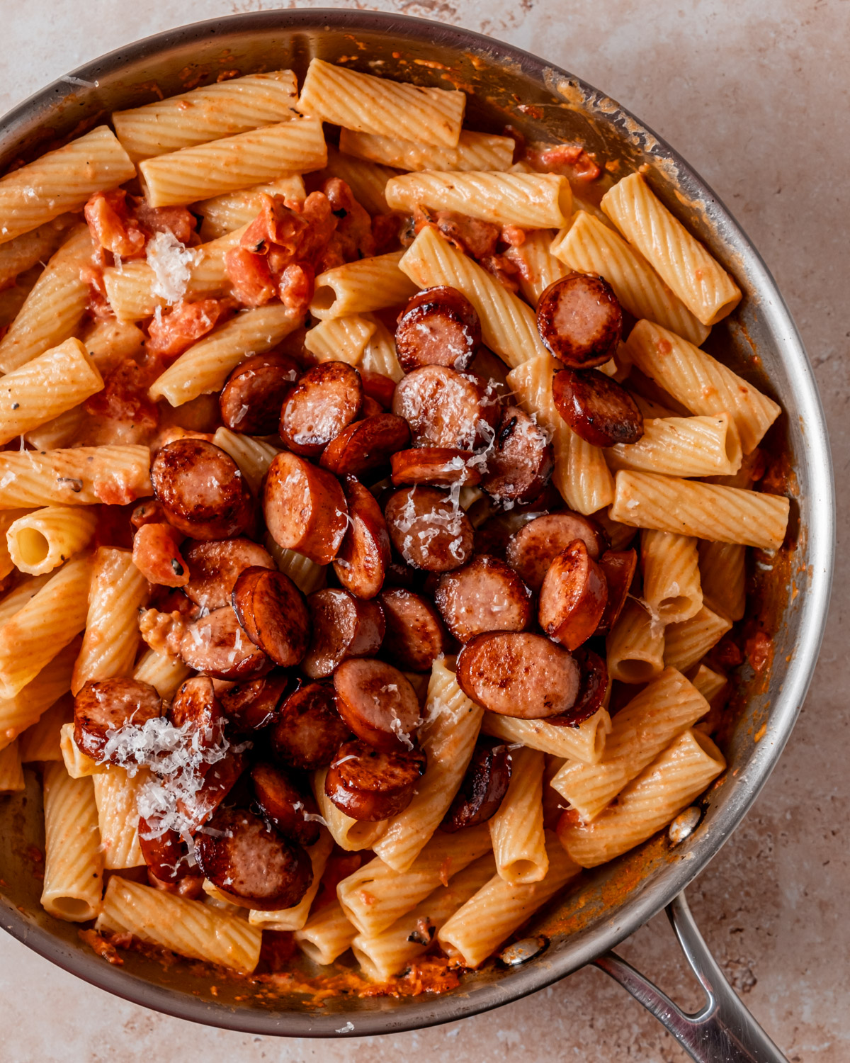 Cooked rigatoni pasta is mixed with browned sausage and creamy tomato sauce in a skillet, topped with grated Parmesan.