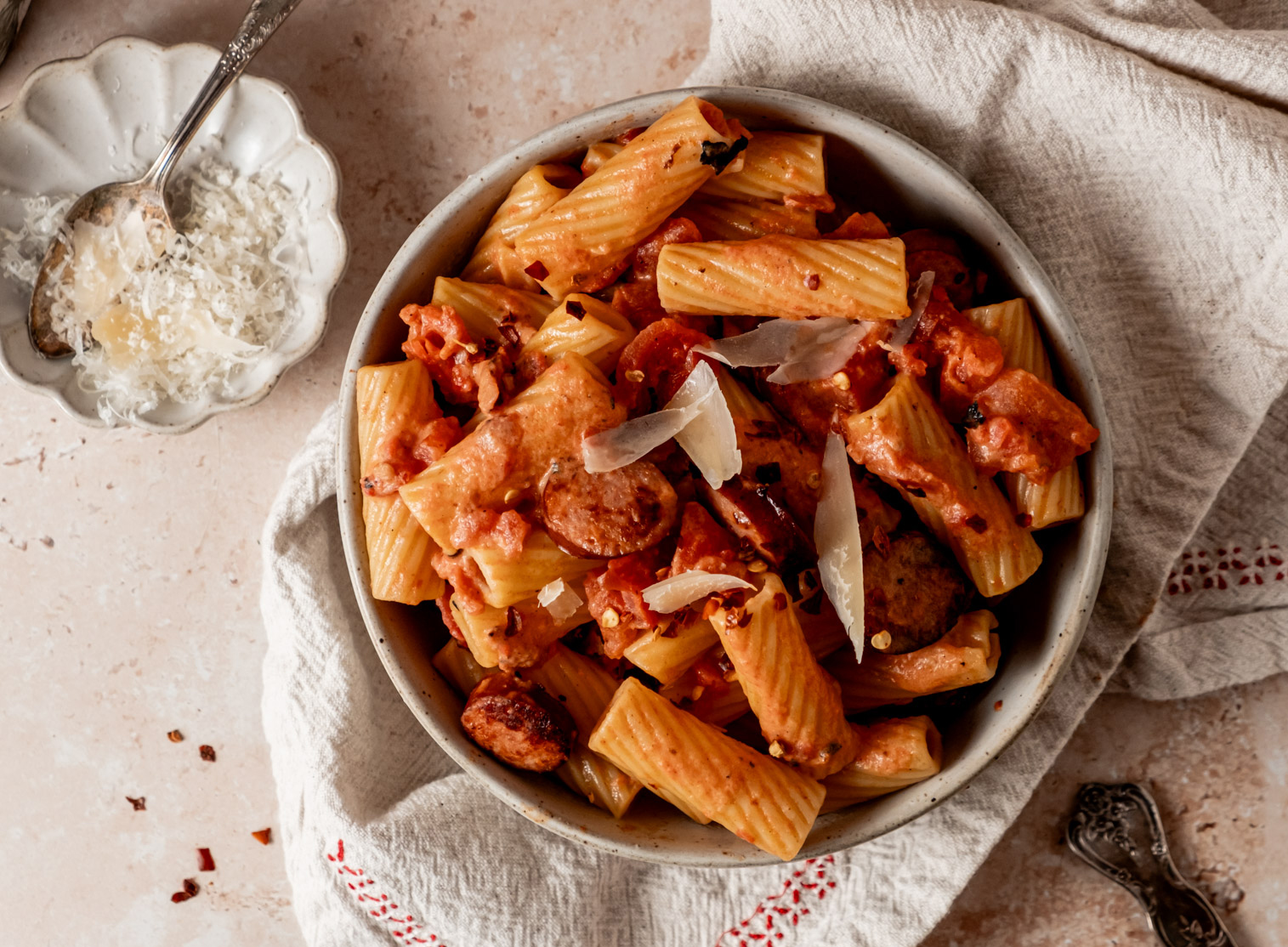 Overhead view of creamy sausage pasta topped with paprika flakes and shaved Parmesan, with a small dish of grated Parmesan beside it.