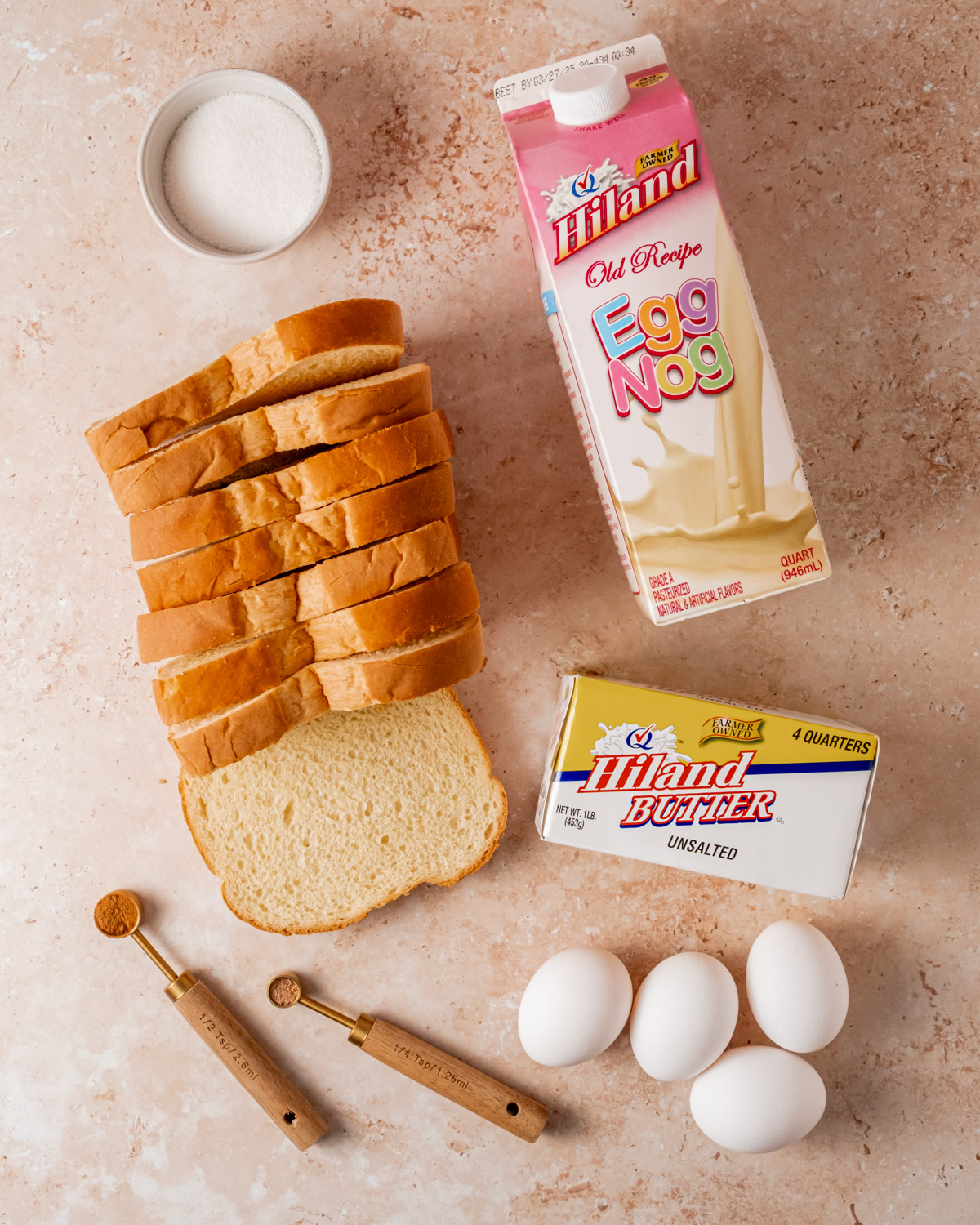 Ingredients for eggnog French toast including a carton of Hiland Egg Nog, a stick of butter, eggs, sugar, spices, and sliced bread on a tan surface.