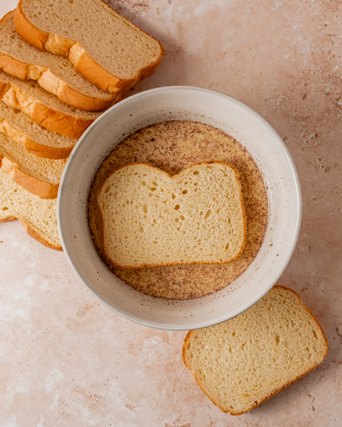 A slice of bread soaking in an eggnog and spice mixture in a mixing bowl, with more slices of bread around the bowl.