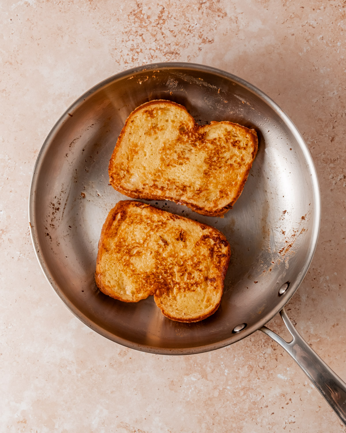 Two slices of French toast being cooked in a stainless steel skillet until golden brown.