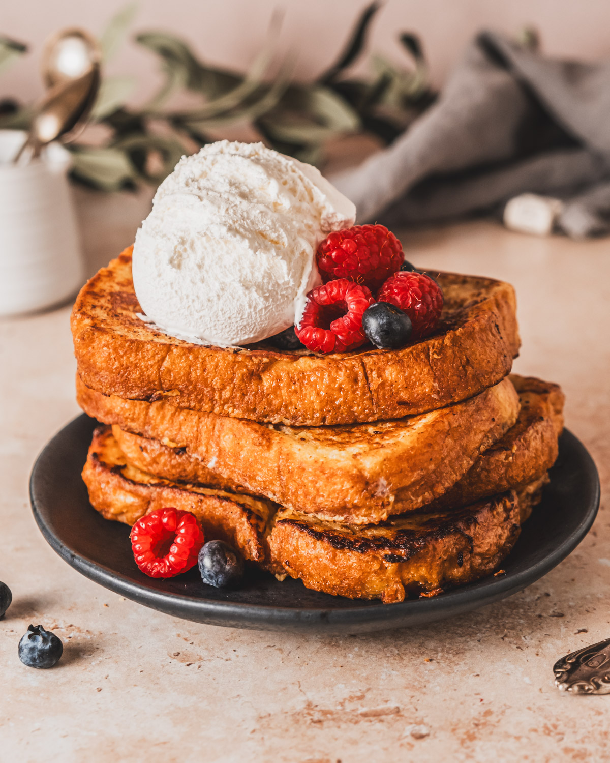 Close-up of stacked French toast slices topped with whipped cream and berries, with a cozy breakfast setting in the background.