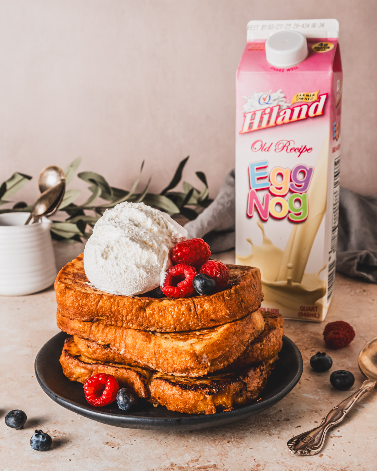 A thick stack of eggnog French toast on a black plate, garnished with a large dollop of whipped cream and fresh berries, with a carton of Hiland Egg Nog in the background.