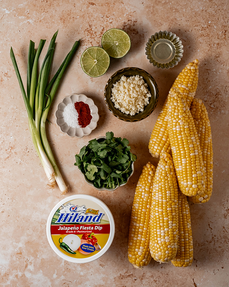 Overhead view of fresh corn, green onions, cilantro, cotija cheese, lime, seasonings, oil, and Hiland Jalapeño Fiesta Dip arranged as ingredients for jalapeño corn salad.
