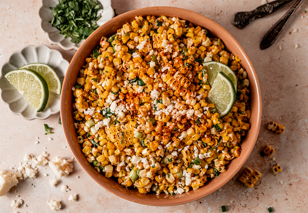 Close-up of a bowl of grilled corn salad mixed with creamy dressing, cilantro, cotija cheese, chili seasoning, and lime wedges, garnished with charred corn and fresh herbs.