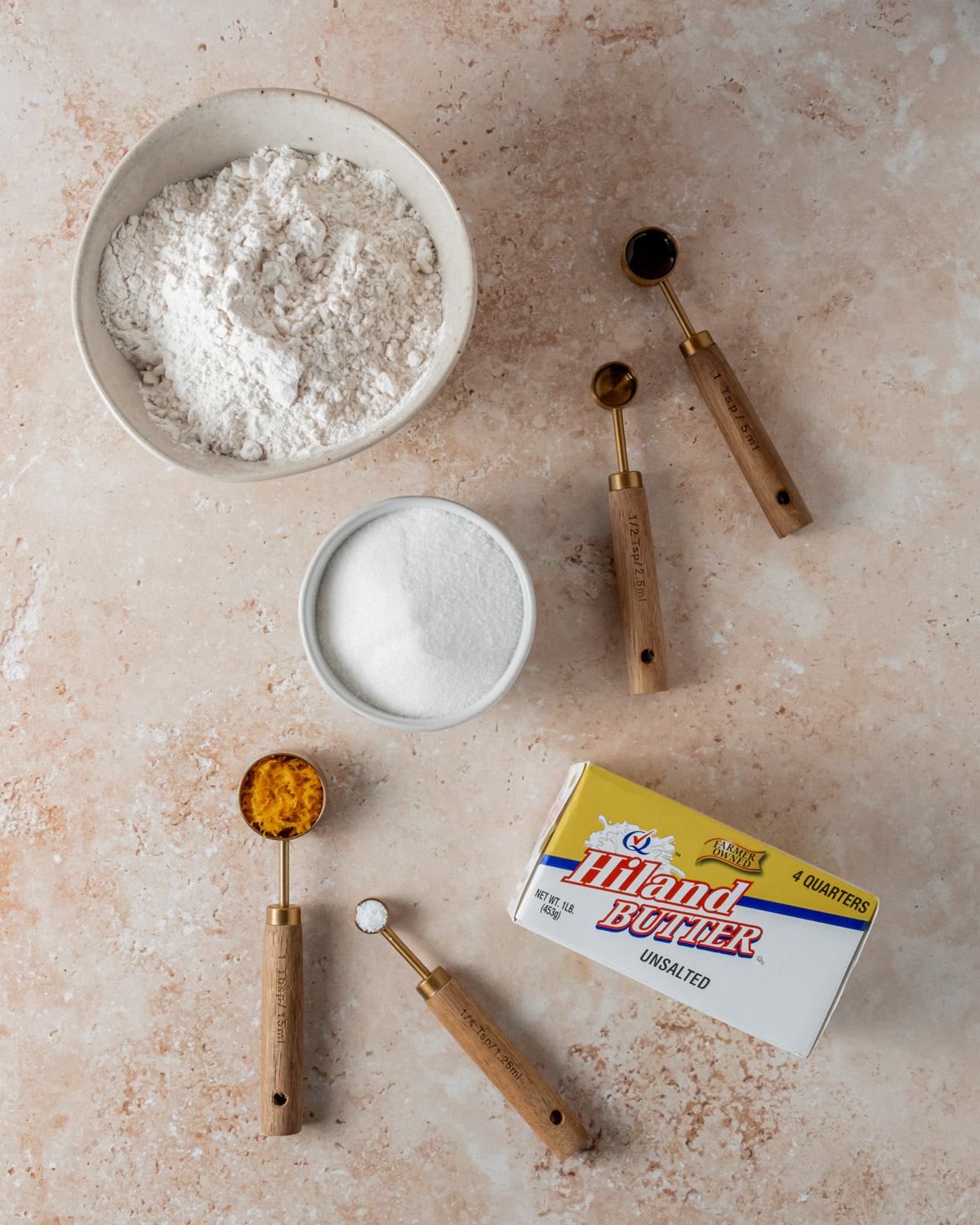 Ingredients for orange shortbread cookies laid out on a beige surface, including flour, sugar, butter, orange zest, and a small dish of salt with wooden measuring spoons.