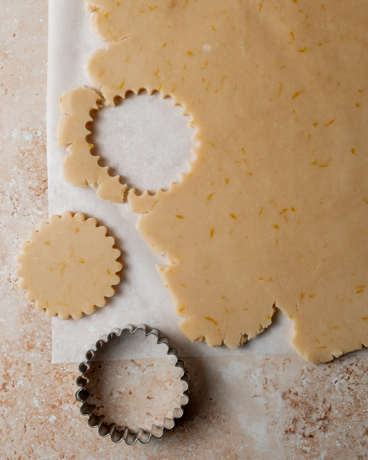 Rolled-out dough with flecks of orange zest, showing a fluted-edge cookie cutter in use, surrounded by neatly cut cookie shapes on a lightly floured surface.