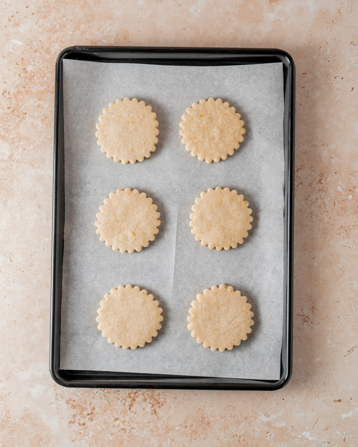 A baking sheet lined with parchment paper featuring six unbaked orange shortbread cookies with fluted edges evenly spaced and ready for the oven.