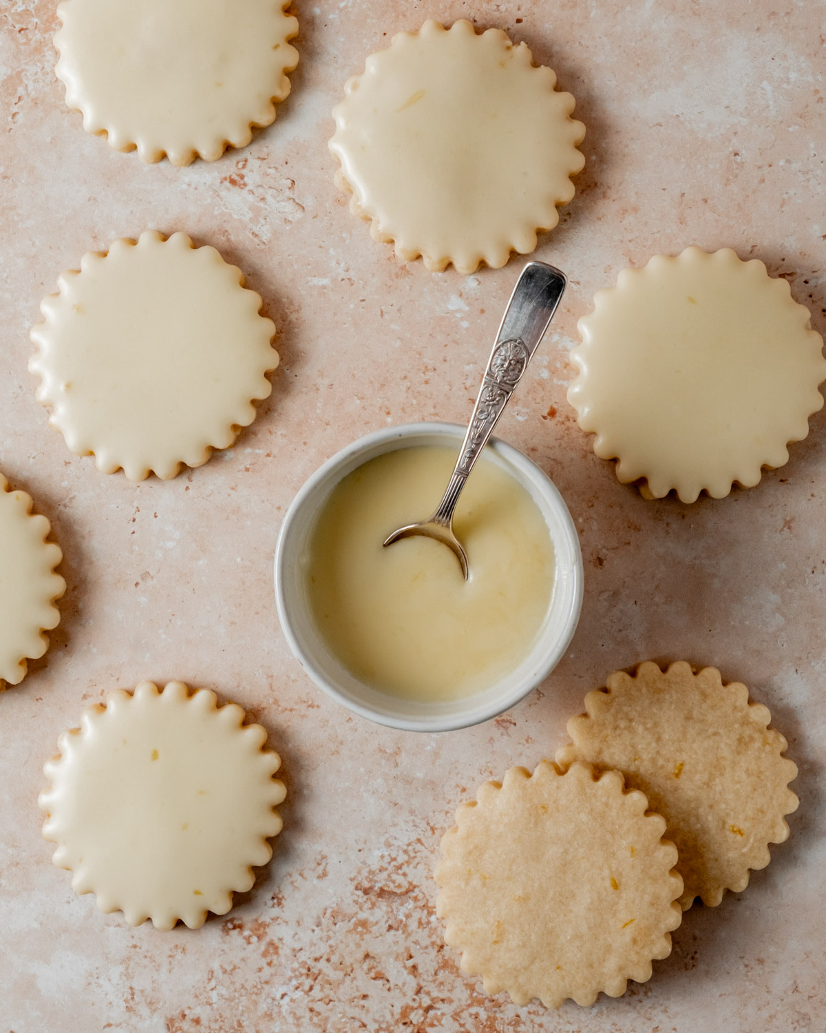 Glazed orange shortbread cookies with a smooth icing finish arranged on a textured surface, with a small bowl of glaze and a spoon.