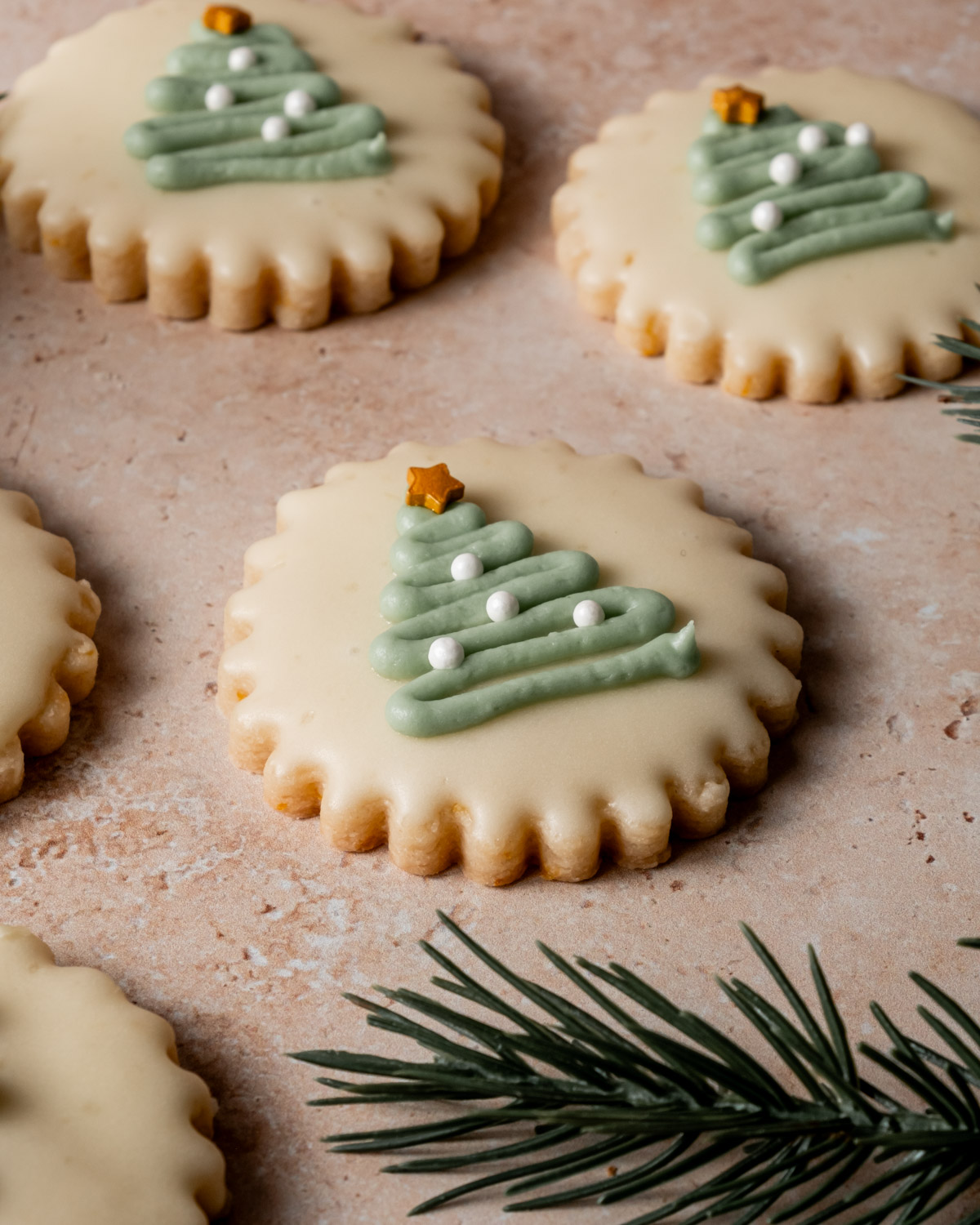 Close-up of a scalloped-edge shortbread cookies with a smooth icing finish, decorated with a green piped frosting Christmas tree design, white pearl-like ornaments, and a golden star at the top, resting on a light beige textured background with pine sprigs.