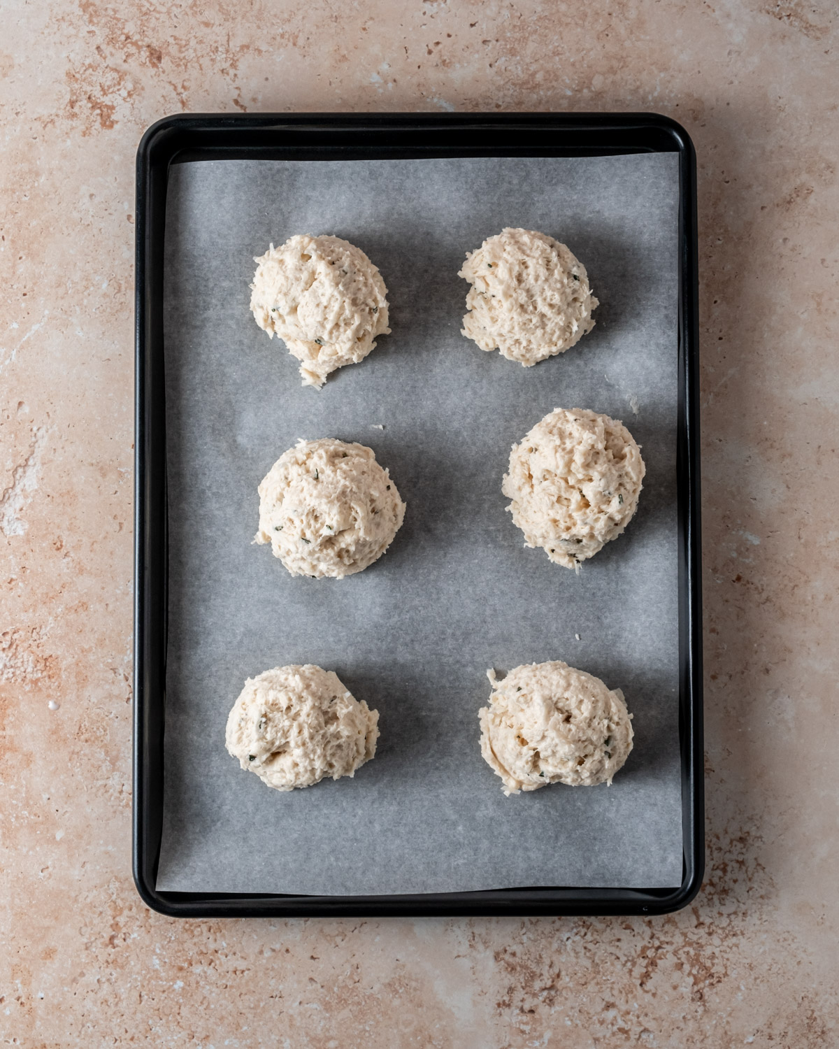 Unbaked Parmesan rosemary biscuit dough scooped onto a parchment-lined baking sheet, ready to go into the oven.