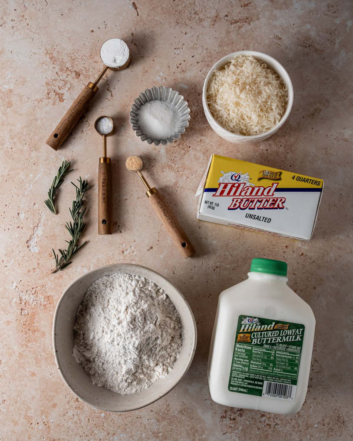 Ingredients for Parmesan rosemary biscuits, including flour, cheese, rosemary, buttermilk, and butter, arranged on a neutral surface.