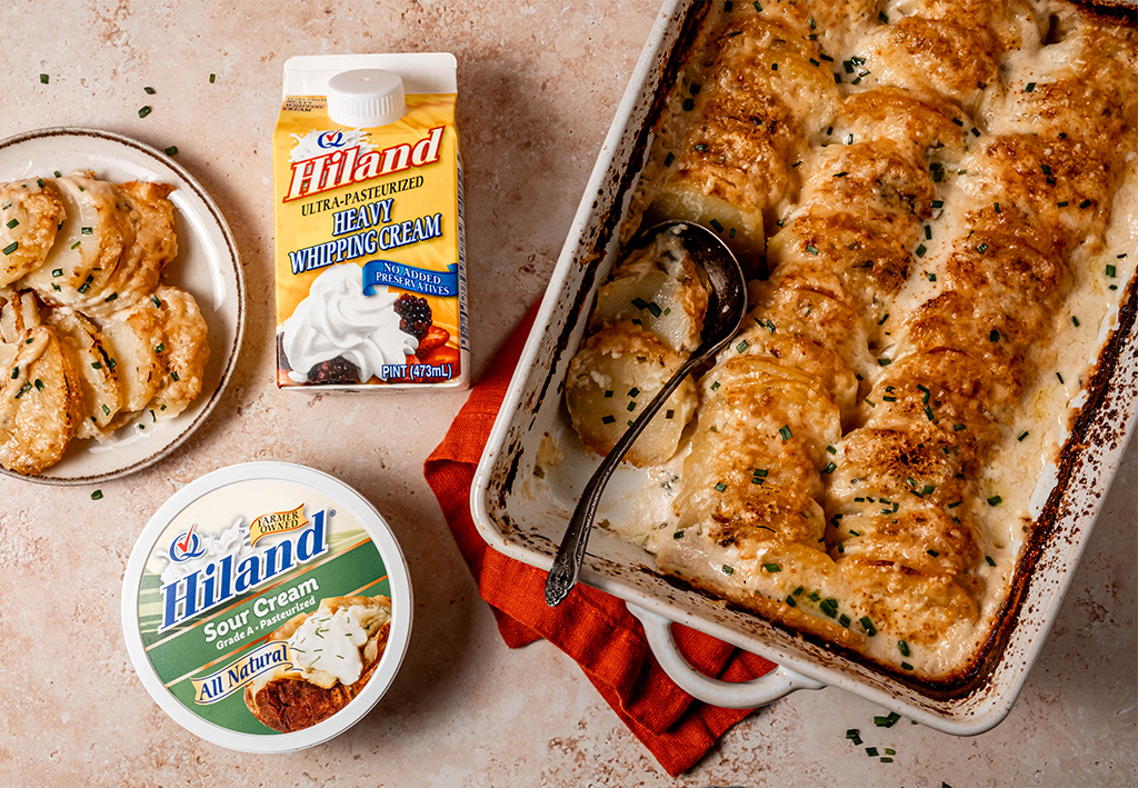 Golden brown sour cream and chive potato gratin baked in a white casserole dish, served beside Hiland Sour Cream and Hiland Heavy Whipping Cream.