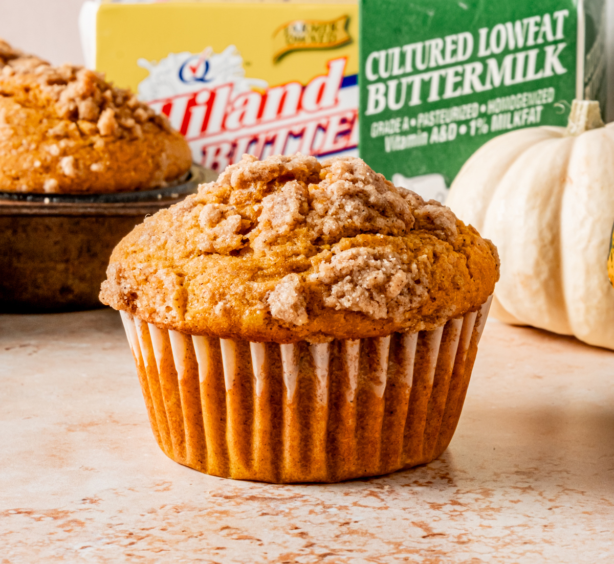 A pumpkin streusel muffin with crumb topping, placed in front of Hiland buttermilk and butter packaging, with pumpkins in the background.